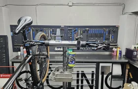 Una bicicleta está montada en un soporte de reparación en el taller de Biciclinic, con varias herramientas organizadas en la pared detrás y suministros de mantenimiento en el mostrador.