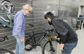 Dos hombres examinan una bicicleta de montaña negra en un taller de Biciclinic con paredes grises, un cuadro de bicicleta en la pared y globos formando el número 29 en el fondo.