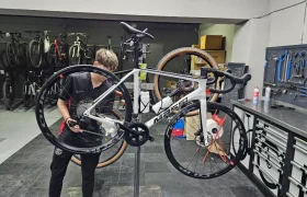 Una persona realiza mantenimiento a una bicicleta de carretera blanca montada en un soporte de reparación en el taller de Biciclinic, con herramientas y otras bicicletas al fondo.