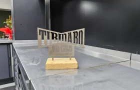 En el entorno industrial del taller de Biciclinic se exhibe un cartel metálico “SUBASTA AL TIBIDABO” con base de madera sobre un banco de trabajo de acero inoxidable.