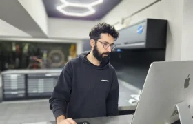 Un hombre con cabello oscuro y barba, que usa anteojos y una sudadera con capucha negra, está de pie frente a un escritorio usando una computadora Apple grande en un espacio de trabajo moderno y bien iluminado.