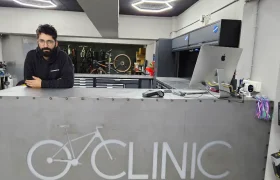 Un hombre está detrás de un mostrador metálico con el gráfico de una bicicleta y la palabra "CLINIC" en Biciclinic, un taller de reparación de bicicletas. Al fondo se ven herramientas, bicicletas y un ordenador.