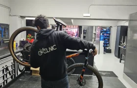 Una persona que viste una sudadera con capucha que dice "CYCLINIC" repara una bicicleta de montaña en un taller de Biciclinic bien organizado, rodeado de herramientas en las paredes y armarios de almacenamiento.