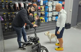 Dos hombres conversan en la tienda de bicicletas Biciclinic; uno sostiene una bicicleta y hace gestos, mientras que el otro permanece con un perro a su lado, frente a una pared de accesorios de ciclismo.