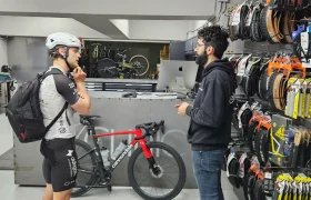 Un ciclista vestido con equipo y casco conversa con un miembro del personal en el mostrador de Biciclinic, mientras en la pared se exhiben neumáticos y accesorios de bicicleta.