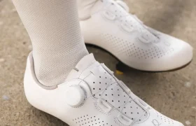 Primer plano de una persona que lleva zapatillas de ciclismo blancas con perforaciones y calcetines blancos, de pie sobre una superficie de hormigón.