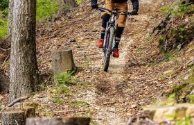 Persona con casco naranja y zapatos rojos monta en bicicleta de montaña por un estrecho sendero de tierra entre árboles altos y follaje verde en el pintoresco bosque Bike Region Glacensis.