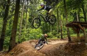 Dos ciclistas de montaña recorren un sendero forestal en la pintoresca Bike Region Glacensis; uno salta desde una rampa mientras el otro toma una curva de tierra más abajo.