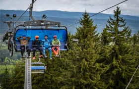 En un día nublado, cuatro personas con casco y ropa de montaña suben a un telesilla azul sobre el paisaje montañoso boscoso de Bike Region Glacensis.