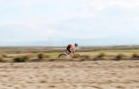Un ciclista con casco y camiseta naranja recorre rápidamente un camino rural en una bicicleta rápida, con un fondo borroso de campos y cielo.