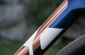 Primer plano del cuadro de la bicicleta BMC Kaius 01 con una combinación de colores cobre metálico y azul con un diseño geométrico blanco, junto a un neumático de bicicleta texturizado, perfecto para cualquiera que busque una bicicleta rápida y actual.