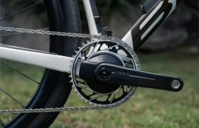 Primer plano de un plato y cadena BMC Kaius 01 en una bicicleta rápida, mostrando el plato, el brazo de la biela y la rueda trasera con hierba al fondo.