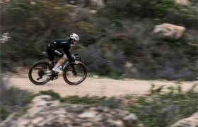 Un ciclista con la equipación del equipo BMC conduce una bicicleta rápida, la BMC Kaius 01, por un camino de tierra a través de un paisaje seco y rocoso con un follaje borroso.