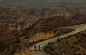 Dos ciclistas recorren un sinuoso camino de tierra a través de un paisaje accidentado y montañoso al anochecer; uno de ellos va en una BMC Kaius 01, disfrutando de la emoción de esta bicicleta rápida bajo un cielo nublado.