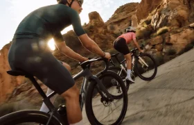 Dos ciclistas con casco y atuendo deportivo montan en bicicletas de carretera BMC Kaius 01 cuesta arriba por una carretera asfaltada a través de un paisaje rocoso e iluminado por el sol.