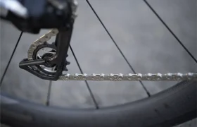 Primer plano de una rueda de bicicleta con una cadena KMC X Flat en el cambio trasero, con parte del neumático visible sobre un fondo gris difuminado.