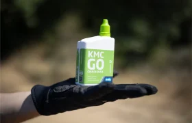 Una mano enguantada sostiene una botella de cera para cadenas KMC GO al aire libre, con un fondo borroso que muestra una cadena de bicicleta KMC X Flat.