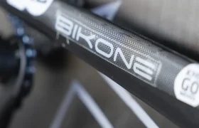 Primer plano de un cuadro de bicicleta rotulado "BIKONE" y "KMC", con cadenas y parte del cambio trasero en foco, que recuerda la precisión vista en las bicicletas del equipo Caja Rural-Seguros RGA.