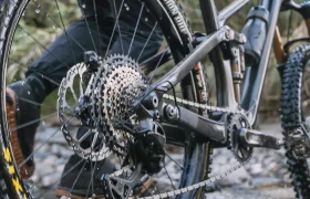 Una persona empuja una bicicleta de montaña equipada con un cambio mecánico Shimano DEORE XT M8200 a través de un arroyo poco profundo, con el agua salpicando alrededor de las ruedas de la bicicleta, lo que garantiza una larga vida útil del cable incluso en condiciones difíciles.