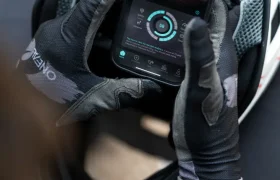 Una persona que lleva guantes negros sostiene un teléfono inteligente que muestra una aplicación de ciclismo, apoyándolo sobre un casco MTB O’Neal Trailfinder Evo blanco y negro.