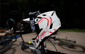 Un casco integral de MTB O’Neal Trailfinder Evo blanco y rojo, equipado con el sistema Quin, descansa sobre el manillar de una bicicleta, con otra bicicleta y una furgoneta al fondo.