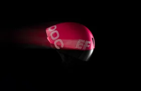 El casco de ciclismo POC Procen Air EF, con un diseño aerodinámico en color rojo y una elegante visera negra, se presenta sobre un fondo oscuro. El efecto de desenfoque de movimiento captura la esencia de la velocidad y el rendimiento.