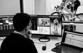 Una persona sentada en un escritorio revisa pantallas de computadora llenas de datos e imágenes de un ciclista con equipo de POC. Al fondo, otra persona pedalea en una bicicleta estática EF Pro Cycling.
