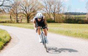 Un ciclista, con casco y gafas de sol, circula en una bicicleta de carretera por una carretera rural con césped verde y árboles sin hojas al fondo, vestido con un maillot Corretto LS de manga larga que le proporciona un rendimiento y una comodidad óptimos.