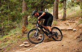 Una persona que lleva casco y ropa negra recorre en bicicleta de montaña, equipada con cubiertas Continental Gravity para MTB, un sendero rocoso y de tierra que atraviesa una zona boscosa, explorando nuevas combinaciones para obtener un rendimiento óptimo.