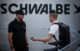Dos hombres de pie delante de un cartel de SCHWALBE; uno sostiene una llave o dispositivo y una bicicleta, mientras que el otro, posiblemente Jordi Bagó de una entrevista Jordi Bagó, lleva una camisa negra y una gorra.