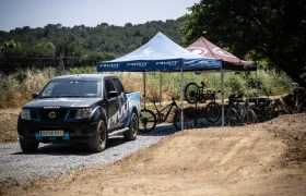 Una camioneta Nissan está aparcada junto a tiendas de campaña en las que se exhiben varias bicicletas y material de ciclismo en un camino de grava cerca de una pista de tierra y árboles, que recuerda a los escenarios de los que habló Jordi Bagó en una entrevista.
