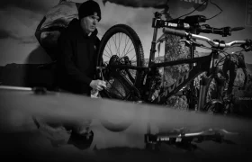 Una persona con gorro y capucha trabaja en el interior de una bicicleta de montaña, limpiando o ajustando la rueda trasera. La escena en blanco y negro parece un momento cándido de una entrevista de Jordi Bagó, que capta el enfoque y la pasión.