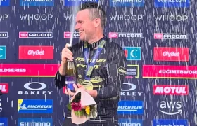 Un piloto de carreras, Jordi Bagó, en traje de carreras, de pie en un podio con una botella en la mano y un micrófono, mientras salpica champán, dando una entrevista Jordi Bagó con logotipos de patrocinadores en el telón de fondo detrás de él.
