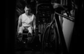 Un hombre con camisa sin mangas utiliza una máquina de extensión de piernas en un gimnasio poco iluminado, con una bicicleta visible en primer plano, que recuerda a un momento cándido de una entrevista Jordi Bagó.