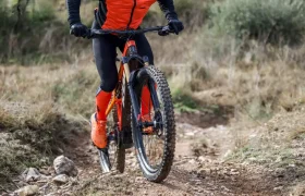 Un ciclista vestido de naranja y negro, inspirado en Entrevista a Josep García, conduce una bicicleta de montaña cuesta arriba por un sendero de tierra rocosa en un entorno rural al aire libre.