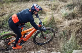 Un ciclista con casco y ropa naranja conduce una bicicleta de montaña por un sendero de tierra a través de un terreno cubierto de hierba, que recuerda escenas de la Entrevista a Josep García.