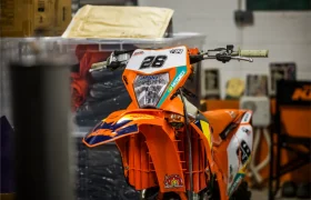 Una moto de cross naranja con el número 26 en la parte delantera está aparcada en el interior cerca de estanterías y cajas de almacenaje, similares a las que se ven en una Entrevista a Josep García.