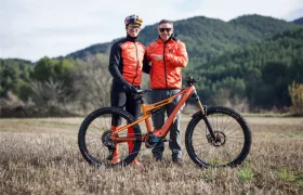 Dos hombres con chaquetas naranjas se paran al aire libre en un campo con una bicicleta de montaña, con árboles y colinas detrás de ellos durante una entrevista a Josep García.