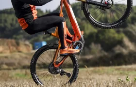 Una persona con casco realiza un caballito en una bicicleta de montaña naranja en un campo abierto con árboles al fondo.