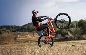 Persona con casco y atuendo naranja realiza un caballito en una bicicleta de montaña naranja en un campo abierto, rodeado de árboles y colinas, que recuerda escenas de una entrevista a Josep García.