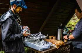 Dos personas preparan y sirven comida y bebida bajo un refugio de madera en Bike Region Glacensis; una sostiene artículos envueltos en papel de aluminio mientras la otra sirve bebidas calientes junto a una bandeja de bollería.