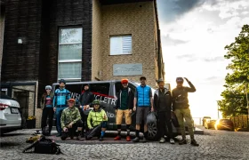 Un grupo de nueve ciclistas posa con su equipo delante de una furgoneta y un moderno edificio al amanecer o al atardecer, listos para explorar las rutas panorámicas de Bike Region Glacensis.