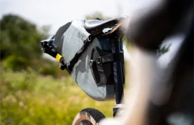 Primer plano de un sillín de bicicleta con una gran bolsa gris EVOC Bikepacking colocada debajo, que ofrece protección adicional, al aire libre con vegetación borrosa de fondo.