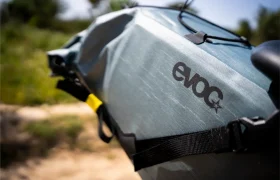 Primer plano de una bolsa para bicicleta de color gris azulado con el logotipo "evoc", sujeta a una bicicleta con correas negras: una pieza esencial del equipo EVOC Bikepacking, perfecta para transportar tu equipo protegido al aire libre en un entorno natural.