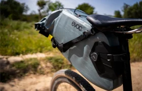 Primer plano de un sillín de bicicleta con una gran bolsa gris EVOC Bikepacking sujeta a la tija del sillín, que ofrece protección adicional, colocado al aire libre en un día soleado con vegetación de fondo.