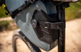 Primer plano de un cuadro de bicicleta con una bolsa de almacenamiento EVOC Bikepacking sujeta por un mecanismo de cierre negro cerca de la tija del sillín, que ofrece protección en sus paseos al aire libre por el camino de tierra.