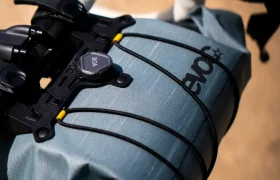 Primer plano de un manillar de bicicleta con una bolsa de manillar EVOC Bikepacking gris, sujeta con cordones elásticos negros y un sistema de esfera BOA para mayor protección.