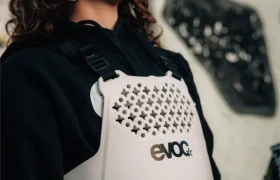 Una persona con una sudadera con capucha negra y una talla XS EVOC Torso Protector blanco se encuentra delante de un mural abstracto, sonriendo.