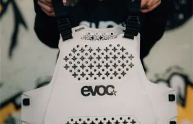 Una persona sostiene un EVOC Torso Protector blanco en talla XS con correas negras, destacando la seguridad, de pie frente a una pared con arte de graffiti negro.