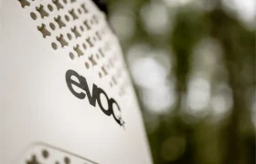 Primer plano de una superficie blanca con orificios de ventilación y el logotipo "evoc" en negro, destacando el EVOC Torso Protector seguridad en talla XS, sobre un fondo verde difuminado.